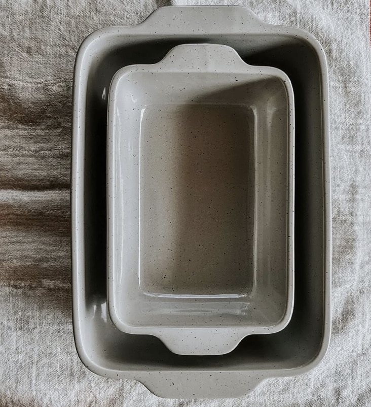 Two gray ceramic baking dishes of different sizes are nested together on a subtle, textured white fabric background. Perfect for your next banket recipe, the dishes have a smooth, glossy finish with speckles and are designed with handles for easy carrying.